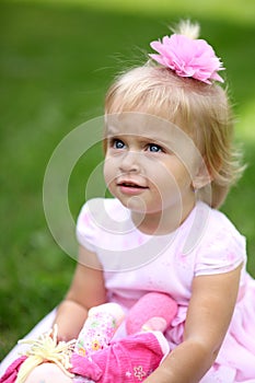 Sweet smiling little girl with long blond hair, sitting on grass in summer park, closeup outdoor portrait.